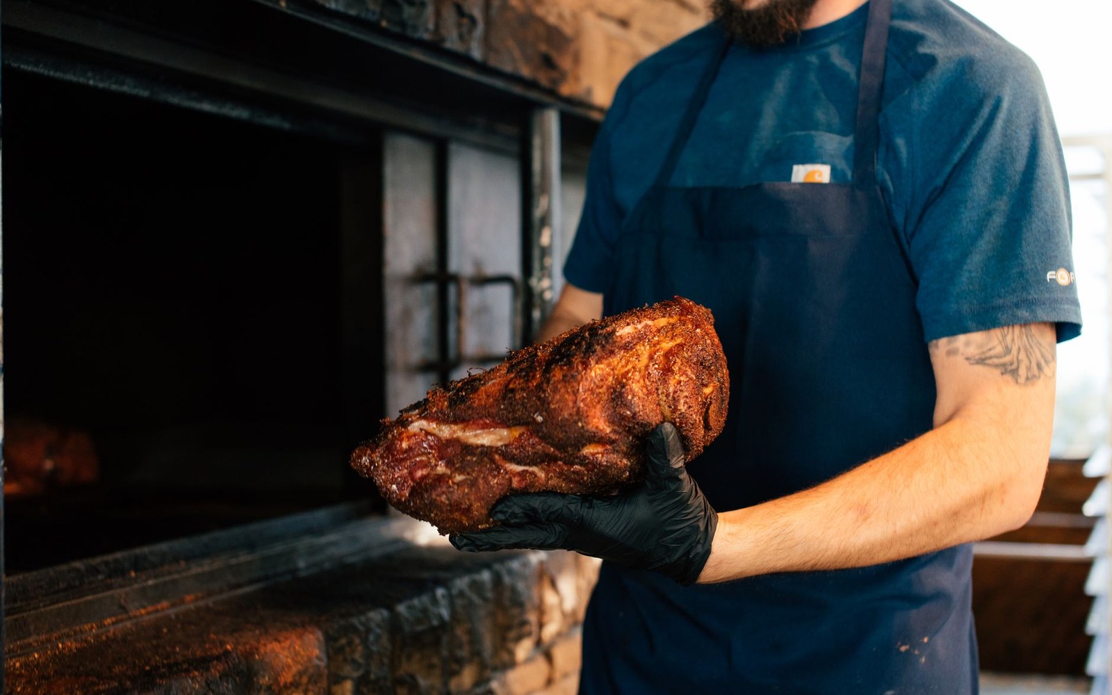 An open pit with pork shoulders on the grate, a pitmaster turning them with tongs, smoke and low light