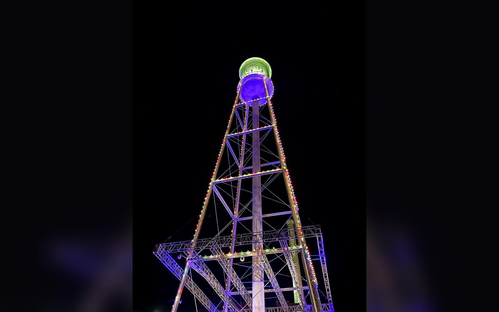 A tall metal water tower structure lit with purple and gold lights against a night sky