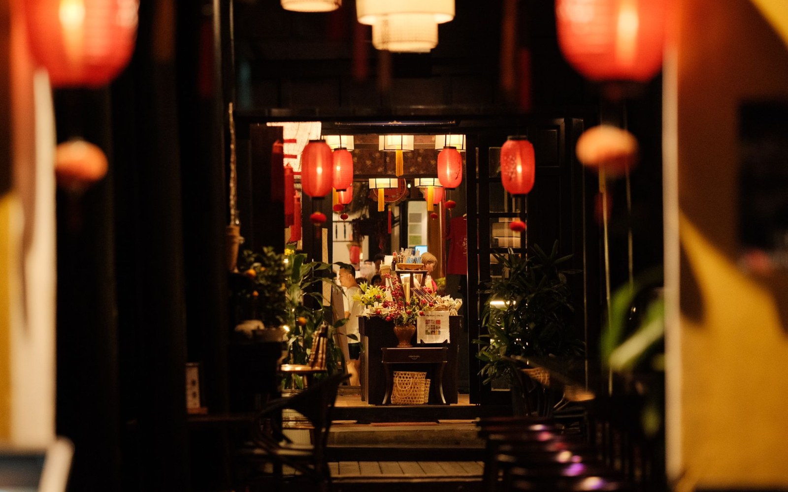Red lanterns hang above a dimly lit restaurant entrance with flowers and potted plants