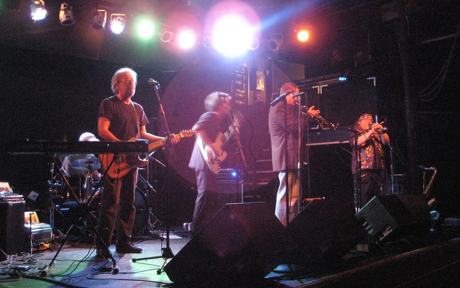 Musicians perform on stage with guitars and a trumpet under colorful stage lighting