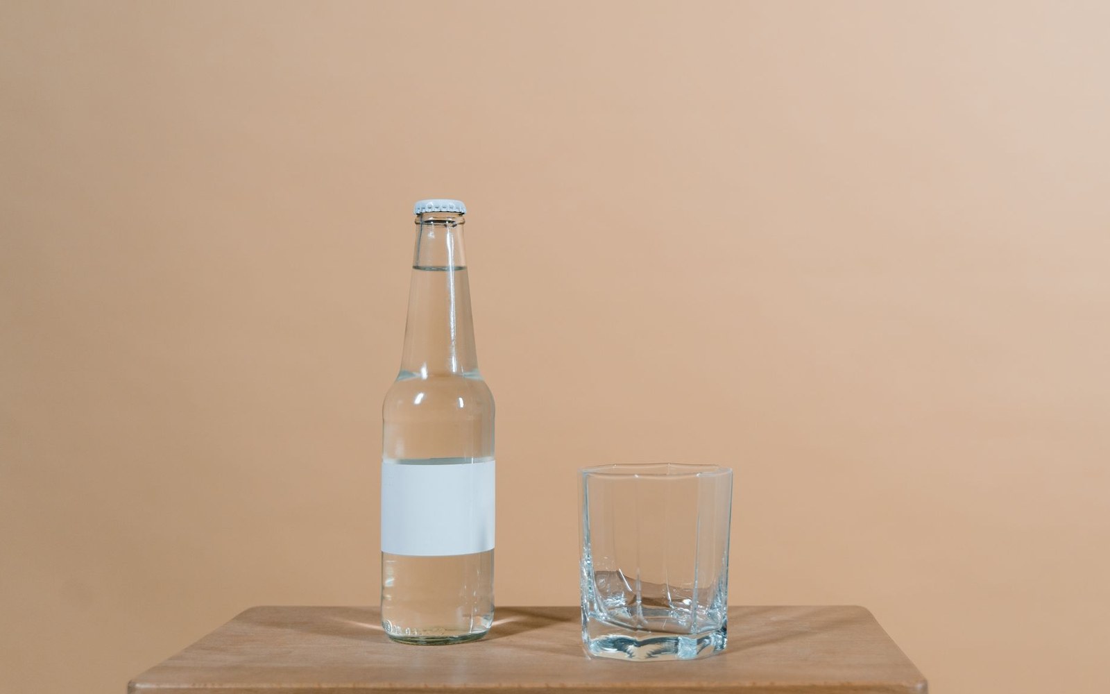 A capped glass bottle with a blank label beside an empty tumbler on a wooden table