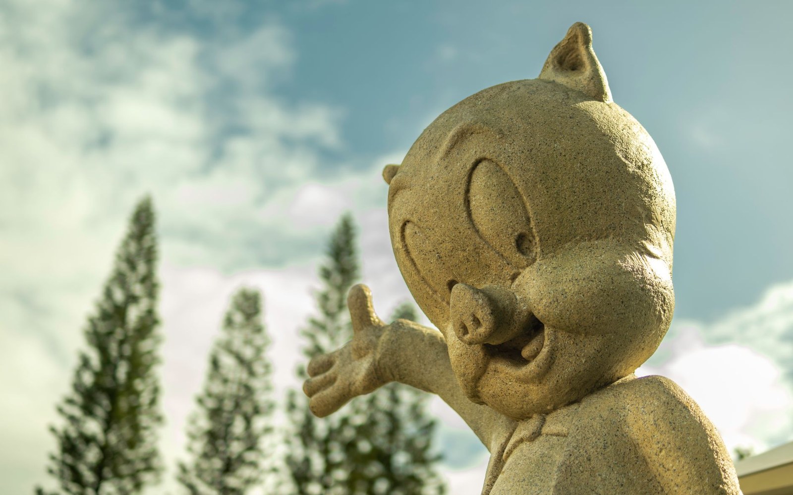 Stone statue of a cartoon pig character raising a hand, against a sky with pine trees