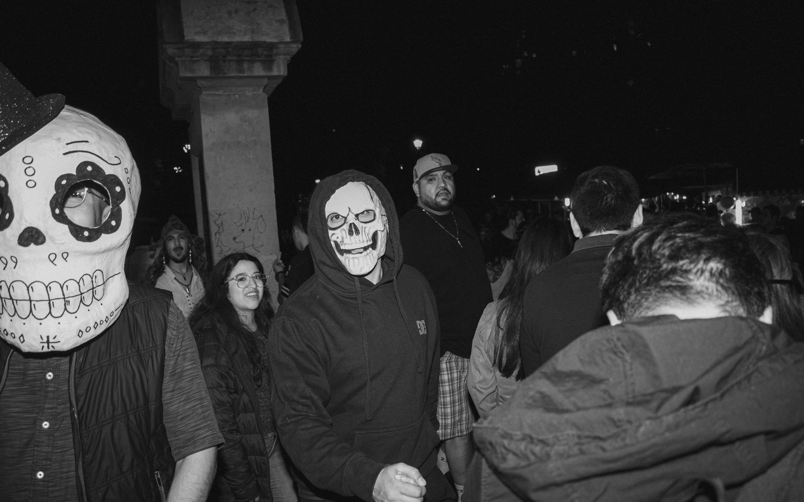 A person wears a hooded skull mask in a nighttime crowd during a street celebration