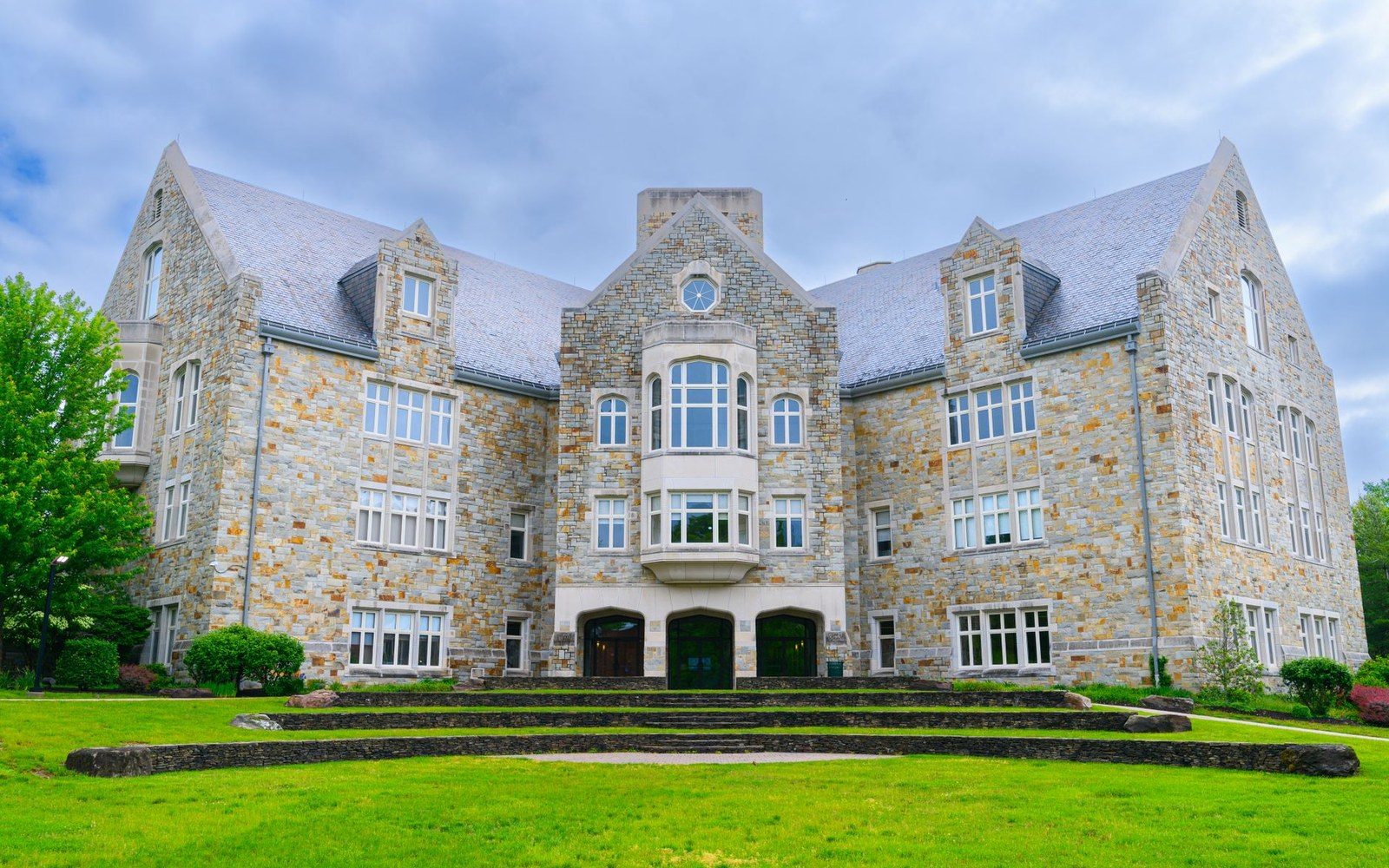 Stone campus building with steep gabled roofs and stairs leading up a grassy lawn