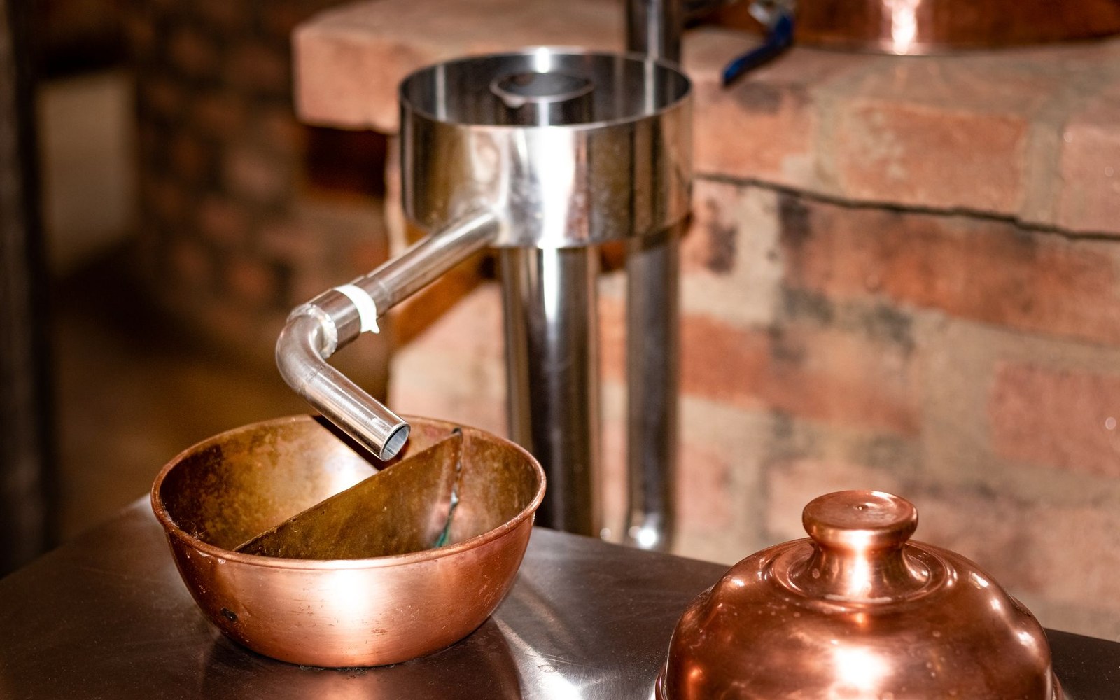 A copper still spout and lid beside a stainless steel condenser in a brick-walled distillery